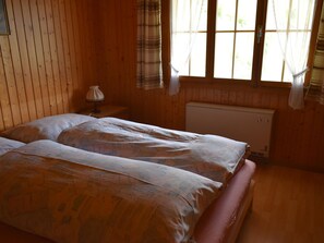 Ein Zimmer mit Holzboden, einem Bett, einem Nachttisch, einem Heizkörper und einem Fenster mit Vorhängen.