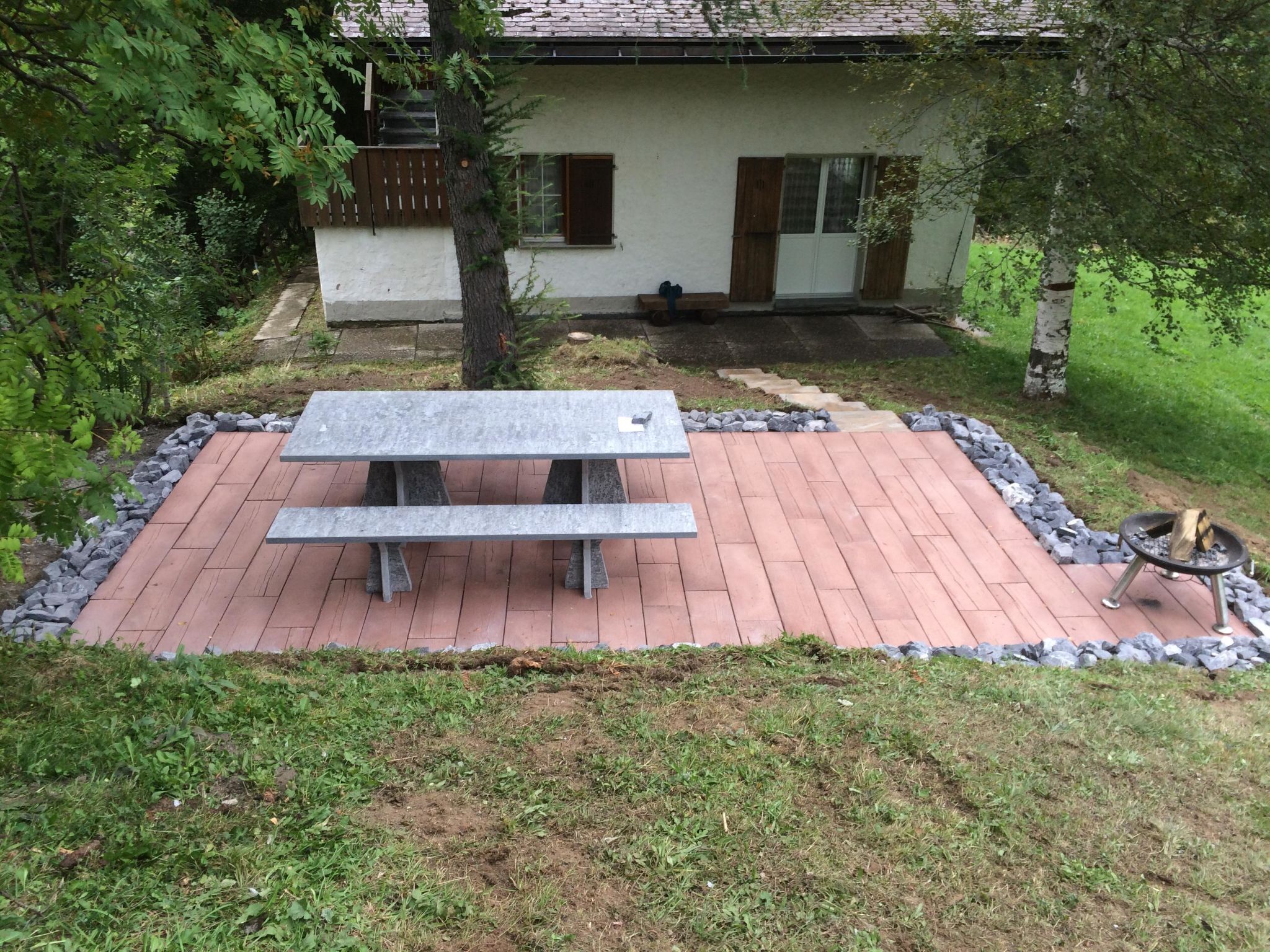 A patio with a stone table and benches, surrounded by a stone border, in front of a house with a white exterior and brown shutters.