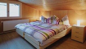 A bedroom with a wooden ceiling, a bed with a striped bedspread, a bedside table with a lamp, and a window with a view of the outside. - Schalmi Appartement/Fewo, Dusche, WC, 2 Schlafräume 3-Zimmerwohnung DG by Interhome (Adelboden)