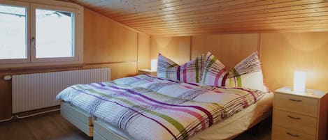 A bedroom with a wooden ceiling, a bed with a striped bedspread, a bedside table with a lamp, and a window with a view of the outside.