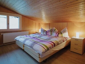 A bedroom with a wooden ceiling, a bed with a striped bedspread, a bedside table with a lamp, and a window with a view of the outside. - Schalmi Appartement/Fewo, Dusche, WC, 2 Schlafräume 3-Zimmerwohnung DG by Interhome (Adelboden)