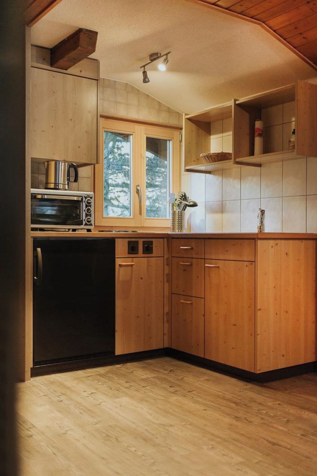 A compact kitchen with wooden cabinets, a built-in oven, and a window with a view of trees.