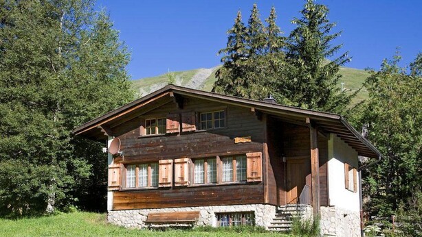 A wooden cabin with a stone foundation, set in a grassy area with trees and mountains in the background. - Schalmi Appartement/Fewo, Dusche, WC, 2 Schlafräume 3-Zimmerwohnung DG by Interhome (Adelboden)