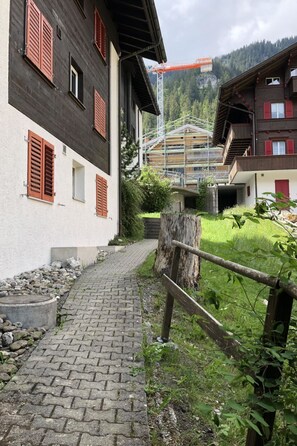 A pathway leading to a building with red shutters, surrounded by greenery and a construction crane in the background. - Sunnegruess UG Appartement/Fewo, Dusche, WC 1.5 Zimmerwohnung by Interhome (Adelboden)