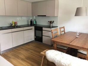 A compact kitchen with a dining area, featuring a wooden table, chairs, and a fluffy white chair. - Sunnegruess UG Appartement/Fewo, Dusche, WC 1.5 Zimmerwohnung by Interhome (Adelboden)