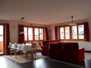 Ein Wohnzimmer mit einem roten Sofa, einem Esstisch mit Stühlen und großen Fenstern.