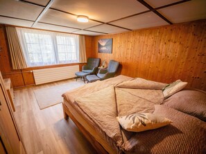 Quarto com cama, duas cadeiras, janela com cortinas, radiador e um quadro na parede.