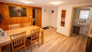 Ruokailutilassa on puinen pöytä ja tuolit, seinällä on kello ja ikkunasta avautuu näkymä keittiöön, jossa on pesuallas ja ikkuna.