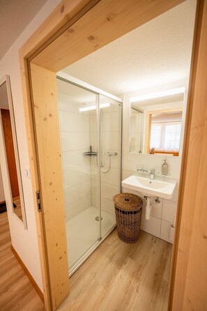 Ein Badezimmer mit Duschkabine aus Glas, einem weißen Waschbecken und einem Korb aus Weidengeflecht.