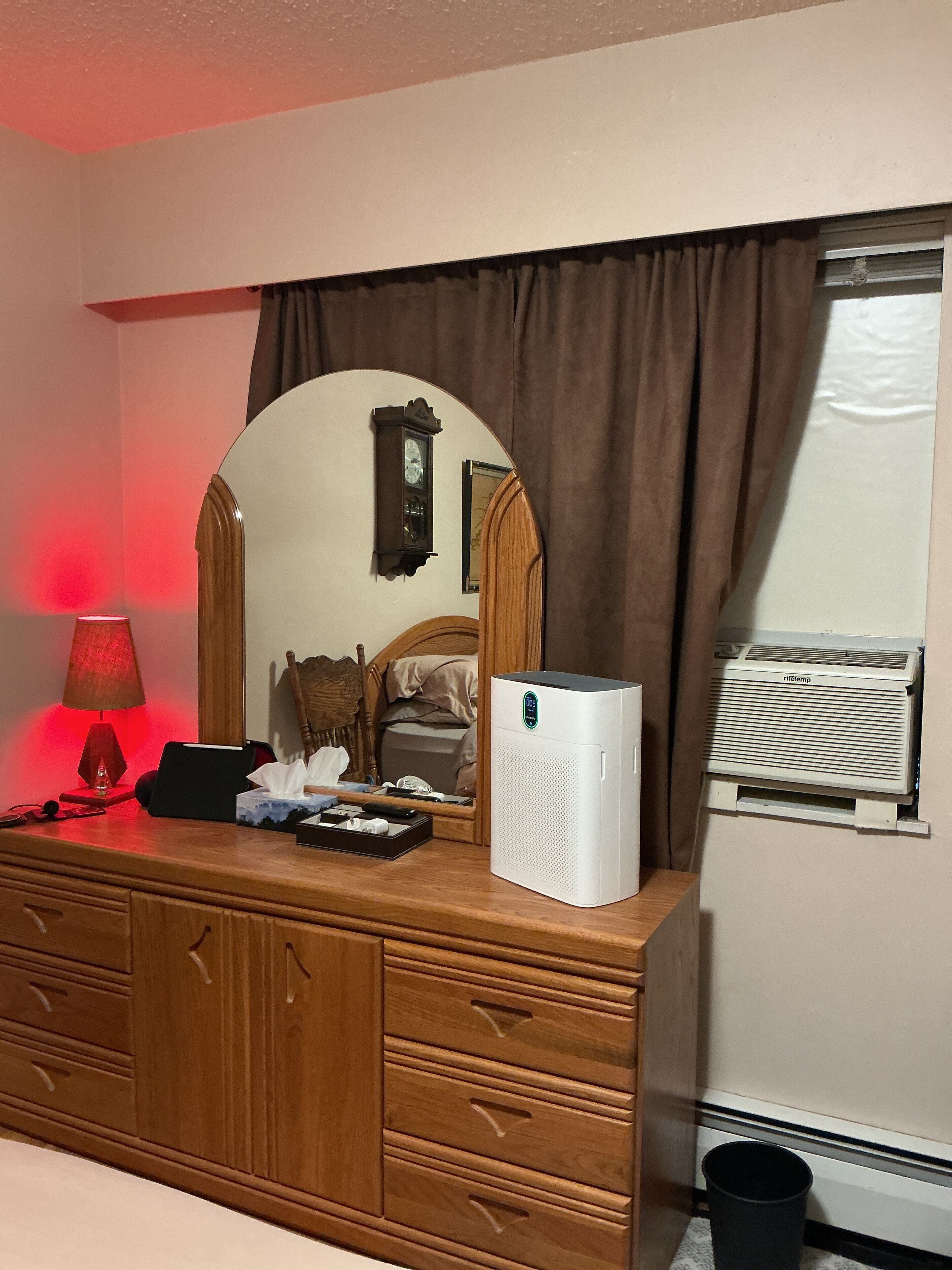A hotel room with a wooden dresser, a mirror, a clock, a bedside table with a lamp, and a window with curtains.