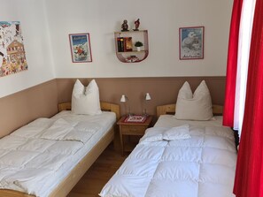 Ein Zimmer mit zwei Einzelbetten, einem kleinen Nachttisch mit Lampe sowie einem Regal mit Büchern und Dekoration.