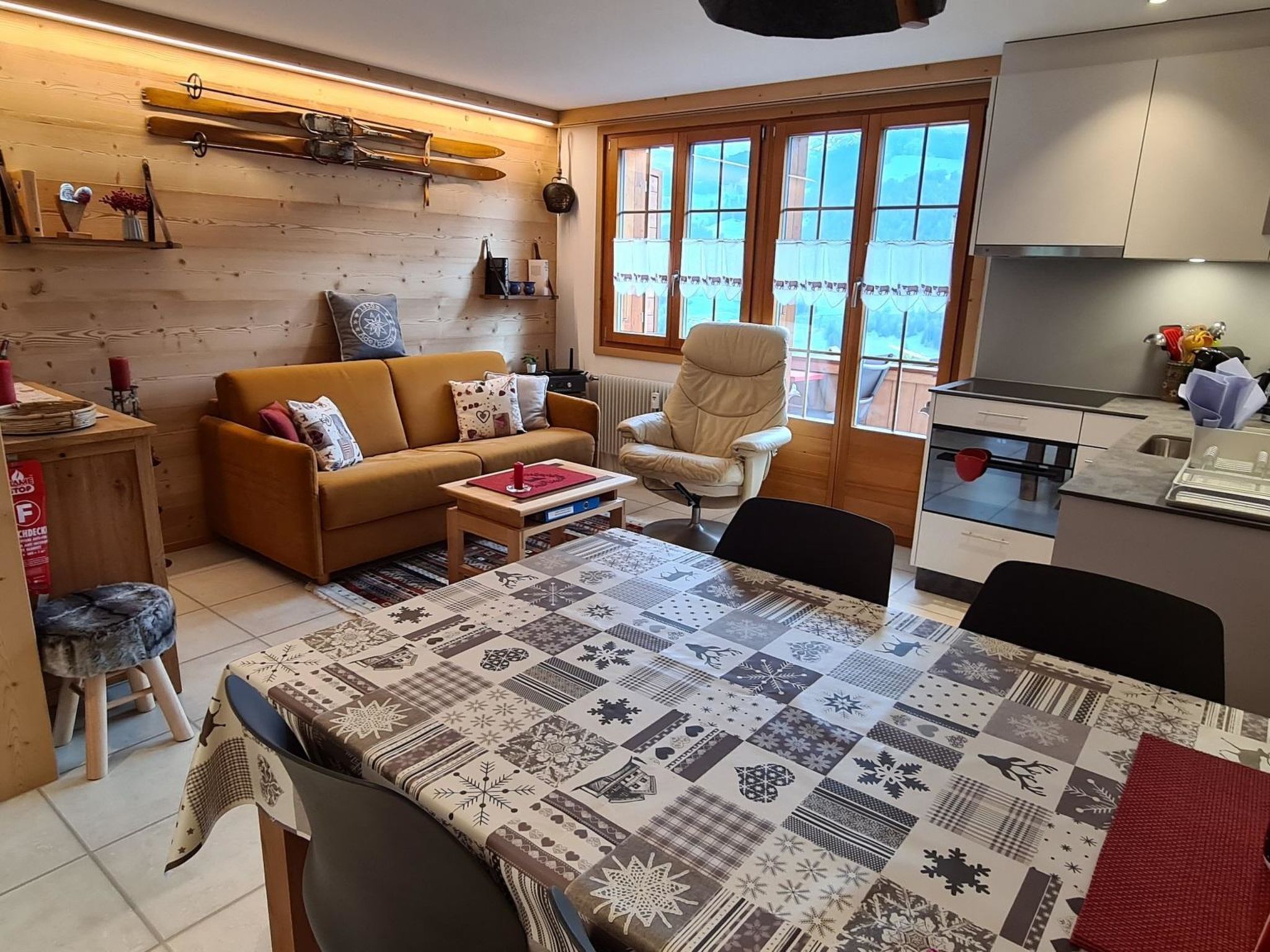 A cozy living room with a sofa, a dining table with a patterned tablecloth, and a kitchen area with cabinets and a window.