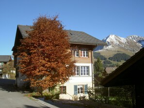 Ein zweistöckiges Gebäude mit Blick auf eine Bergkulisse, ein großer Baum im Vordergrund und ein klarer Himmel.