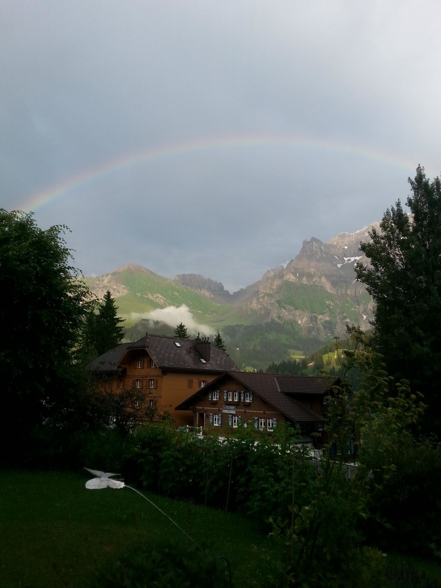 Eine Berglandschaft mit einem Haus im Vordergrund und einem Regenbogen im Hintergrund.