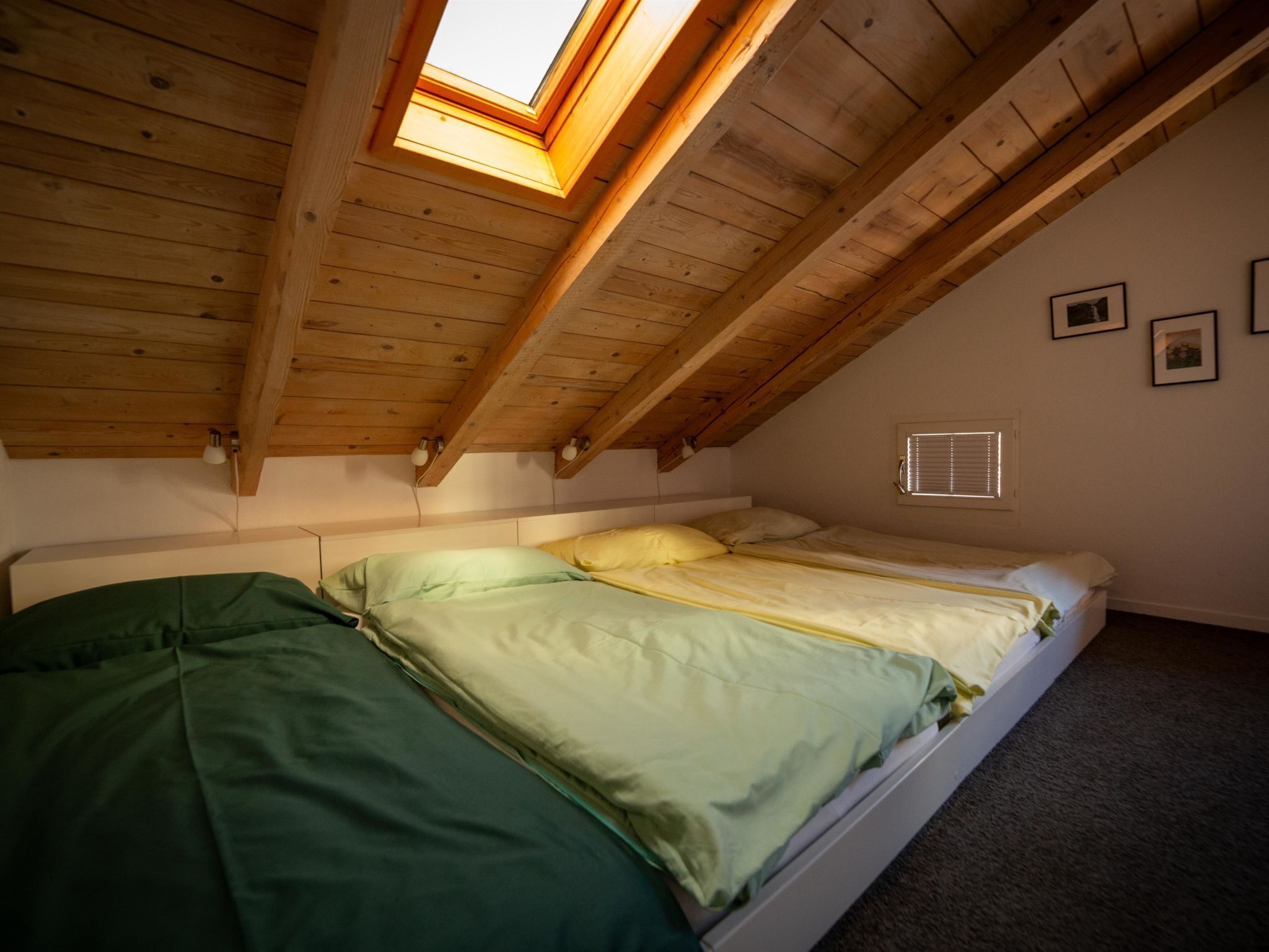 Een slaapkamer met een schuine kap, een dakraam, een bed met groen en geel beddengoed en een klein raam met jaloezieën.