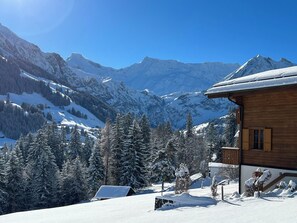 Eine Holzhütte mit schneebedecktem Dach, umgeben von schneebedeckten Bäumen und Bergen im Hintergrund.