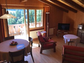Interior de madeira com mesa de jantar, cadeiras, sofá, televisão e varanda com vista para árvores.