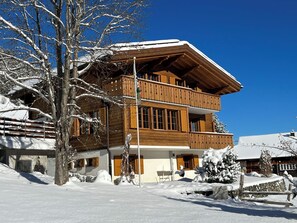 Eine zweistöckige Holzchalet mit Balkon, umgeben von schneebedeckten Bäumen und blauem Himmel.