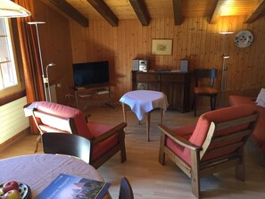Um quarto acolhedor com paredes de madeira, uma televisão, uma escrivaninha com computador e duas cadeiras com almofadas vermelhas.