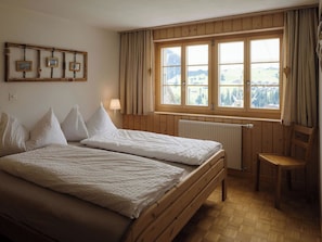 Ein Zimmer mit zwei Betten, einem Holzstuhl, einem Fenster mit Vorhängen und einer Heizung.