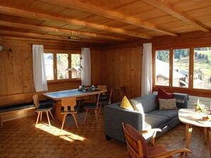 Ein Holzinterieur mit Esstisch, Stühlen, einer Couch und großen Fenstern mit Blick ins Freie.