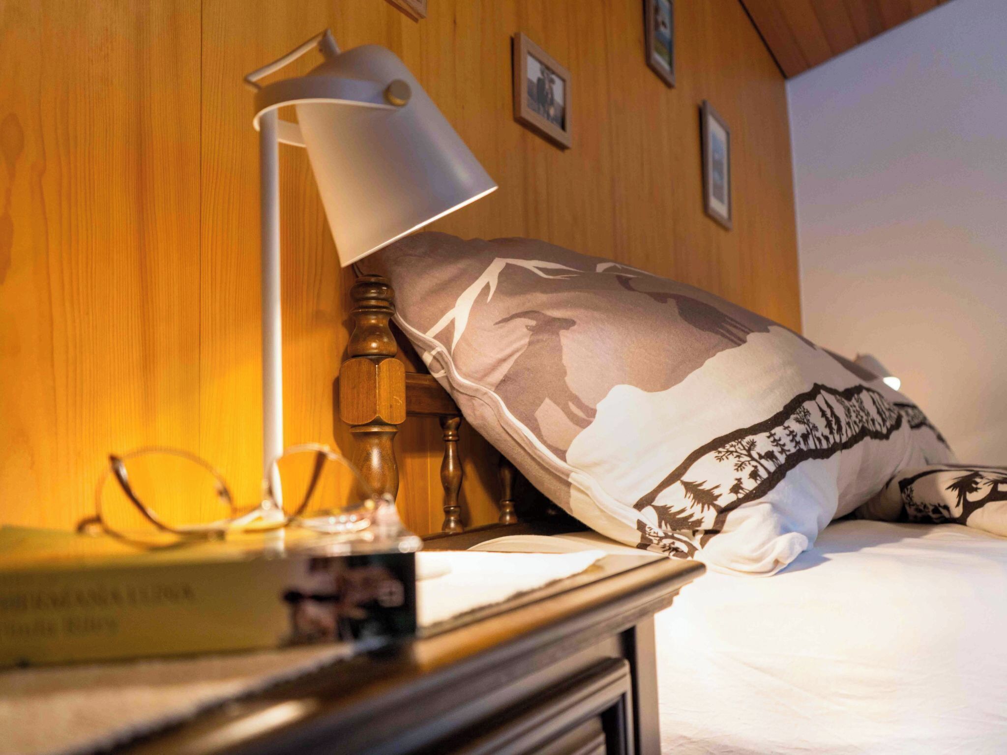 Een net opgemaakt bed met een sierkussen met een natuurprint, een bedlampje, een boek en een paar brillen op een houten nachtkastje.