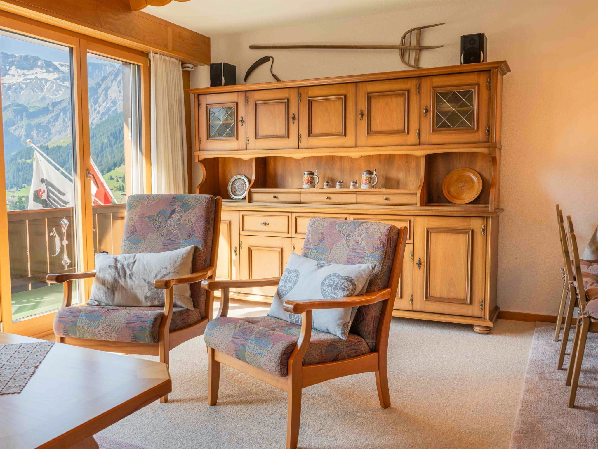 Ein gemütliches Zimmer mit zwei Holzsesseln, einem Schrank mit Glastüren und Blick auf Berge durch große Fenster.