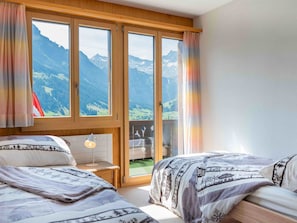 A bedroom with two beds, a nightstand, and a view of mountains through large windows. - Ravinel 2 Appartement/Fewo, Dusche, WC, 3 Schlafräume 4-Zimmerwohnung by Interhome (Adelboden)