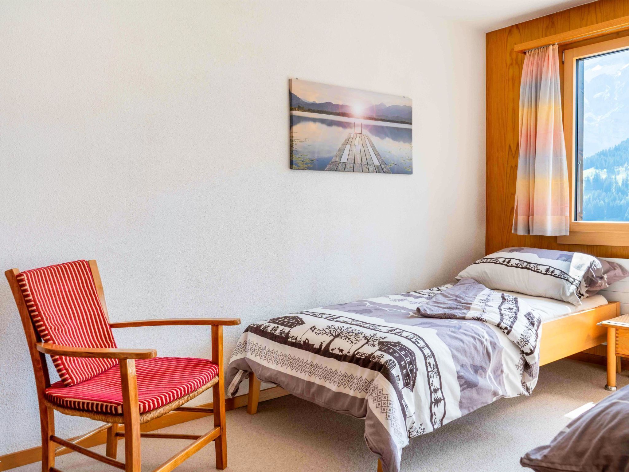 Une chambre à coucher avec un lit simple, une chaise en bois avec un coussin rouge, un tableau représentant un paysage et une fenêtre avec des rideaux.