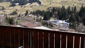 A balcony with a table, chairs, a bottle of water, and a glass. In the background, there are snow-covered mountains and a valley with houses and trees. - Adlerhorst Appartement/Fewo, Dusche, WC, 2 Schlafräume 3-Zimmerwohnung by Interhome (Adelboden)