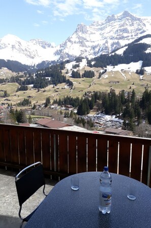Ein Balkon mit Tisch, Stühlen, einer Wasserflasche und einem Glas. Im Hintergrund verschneite Berge und ein Tal mit Häusern und Bäumen.