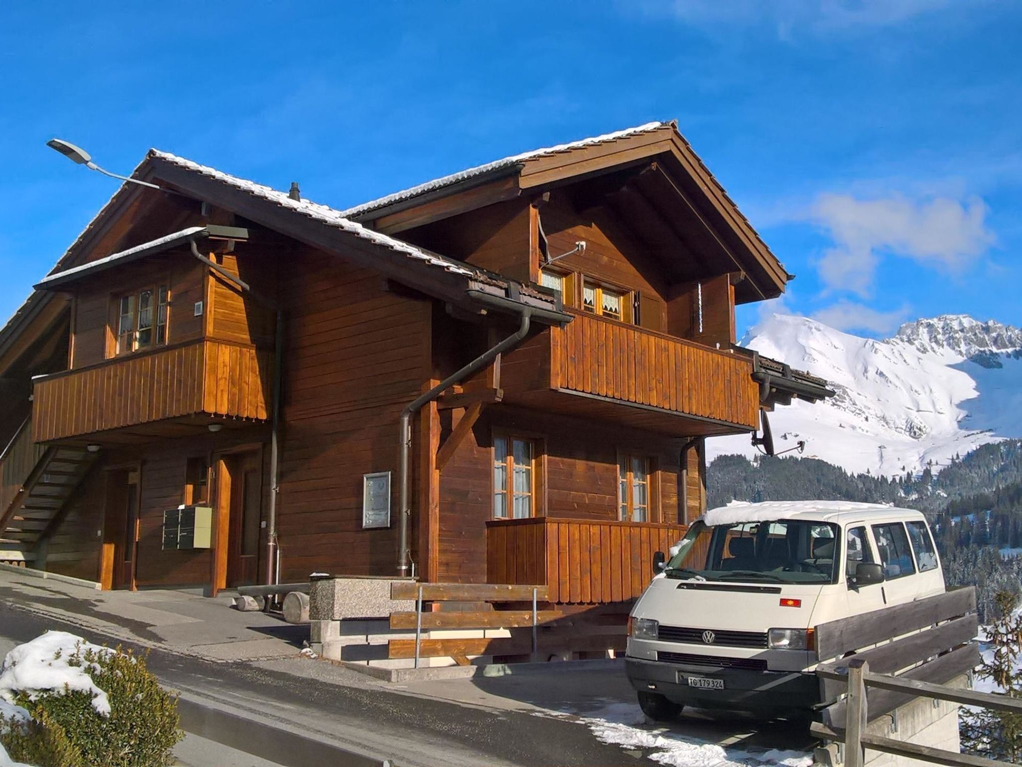 A wooden chalet with a balcony, a staircase, and a parked van in front.