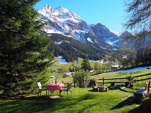 Eine gebirgige Landschaft mit freiem Blick auf schneebedeckte Gipfel, einer Wiese mit Picknicktisch und Stühlen sowie einem kleinen Gewässer im Tal.