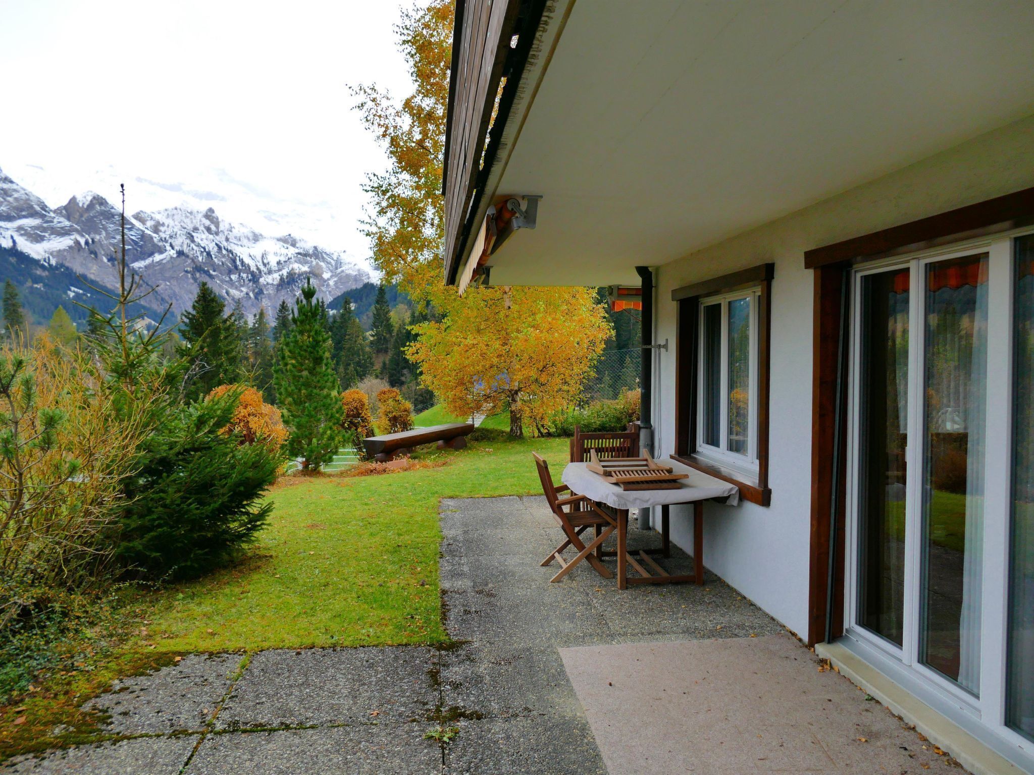 A patio area with a table and chairs, a small garden with grass and shrubs, and a view of mountains in the background.