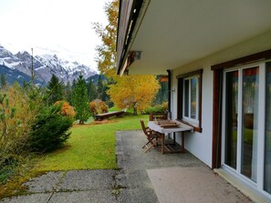 Uma área de pátio com mesa e cadeiras, um pequeno jardim com grama e arbustos, e vista para as montanhas ao fundo.