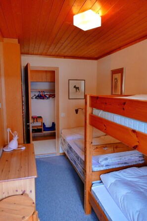 Um quarto com uma cama beliche, uma escrivaninha, uma cadeira, um guarda-roupa e uma porta que dá para outro cômodo.