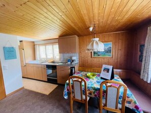 Eine kleine Küche mit Holzausbau, einem Esstisch mit Stühlen, einer Hängeleuchte und einem Bild an der Wand.