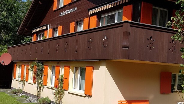 Un chalet con fachada de madera, varias ventanas con contraventanas y un sendero de piedra que lleva a un banco.