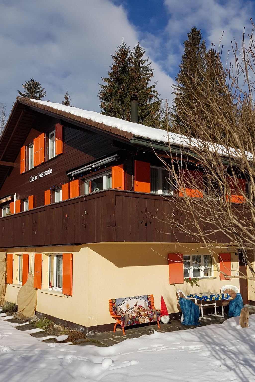 Un chalet dont le toit est recouvert de neige, une enseigne indiquant « Chalet Rosmarie », et une table décorée sur le thème de Noël à l’extérieur.