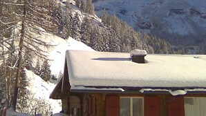 Una cabaña de madera con techo nevado, rodeada de árboles y montañas cubiertas de nieve.