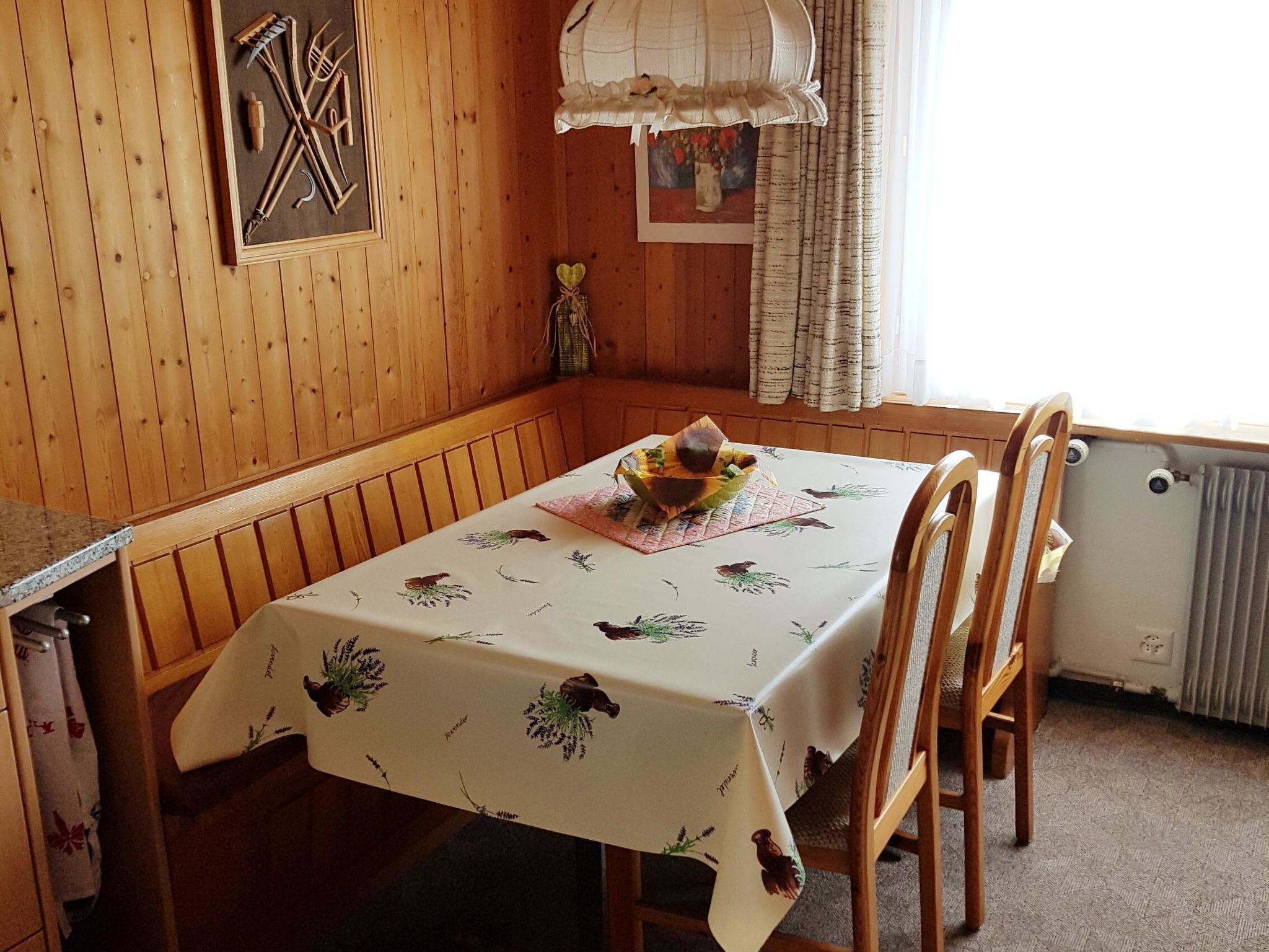 Une salle à manger en bois avec une table recouverte d’une nappe à motifs floraux, des chaises en bois, une fenêtre avec des rideaux et un radiateur.
