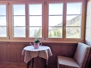 A room with a wooden interior, a table with a tablecloth, a chair, and a window with a view of a mountainous landscape.