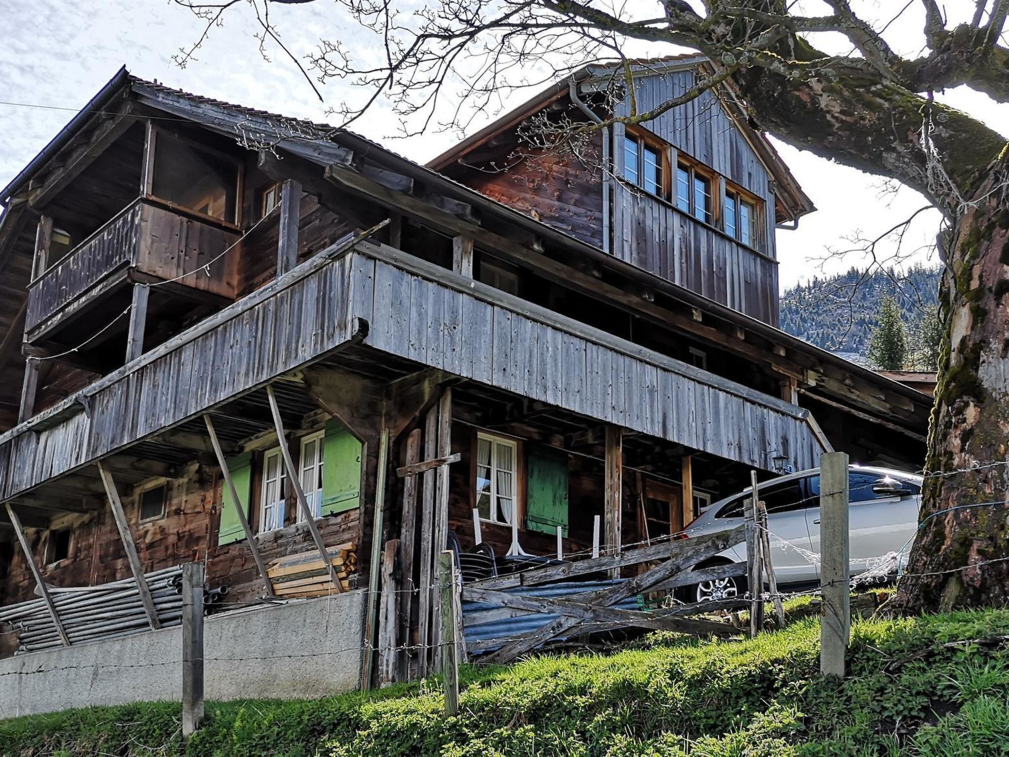 Une maison en bois à deux étages, dotée d’un balcon, avec une voiture garée devant et un arbre sur la droite.