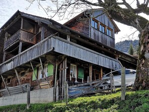 A two-story wooden house with a balcony, a car parked in front, and a tree to the right.