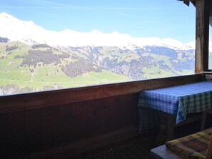 A wooden cabin with a table and bench, overlooking a mountainous landscape with snow-capped peaks and green valleys.