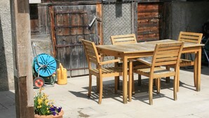 Eine Terrasse im Freien mit einem Holztisch und -stühlen, einem Blumentopf und einem blauen Gartenschlauch.