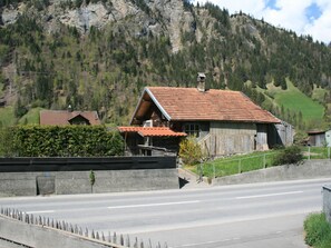 Ein ländliches Haus mit geneigtem Dach, einer Steinmauer im Vordergrund und einer Straße.