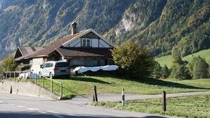 Ein traditionelles Chalet mit geneigtem Dach, einem parkenden Auto und einer Bergkulisse.