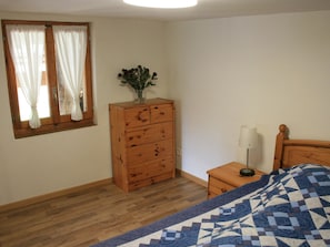 Ein Schlafzimmer mit einem hölzernen Schminktisch, einem Nachttisch mit Lampe, einem Bett mit gemustertem Überwurf und einem Fenster mit Vorhängen.