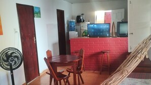 Una habitación con una mesa de comedor, sillas, una hamaca, una cama con manta a rayas, un ventilador y una kitchenette con pared de ladrillo rojo.